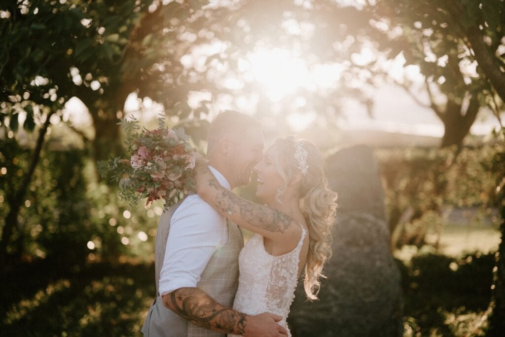 In the heart of Alfriston Gardens, a couple embraces joyfully, with the bride holding a bouquet. Warm sunlight filters through the trees, creating a romantic and serene wedding atmosphere. Image by Pearce Wedding Photography.