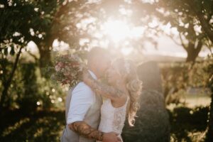 In the heart of Alfriston Gardens, a couple embraces joyfully, with the bride holding a bouquet. Warm sunlight filters through the trees, creating a romantic and serene wedding atmosphere. Image by Pearce Wedding Photography.