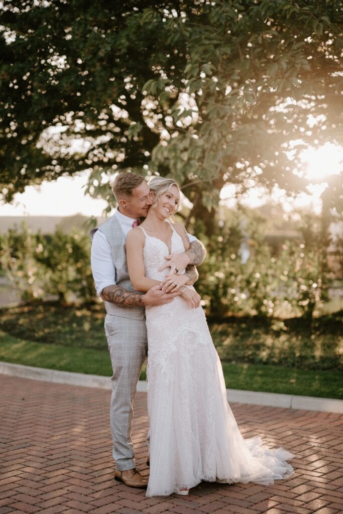 In the enchanting setting of Alfriston Gardens, a couple embraces amidst a sunlit sanctuary. The man in a gray suit holds the woman in her white lace wedding dress as they share warm smiles, surrounded by lush greenery and soft sunlight filtering through the trees. Image by Pearce Wedding Photography.