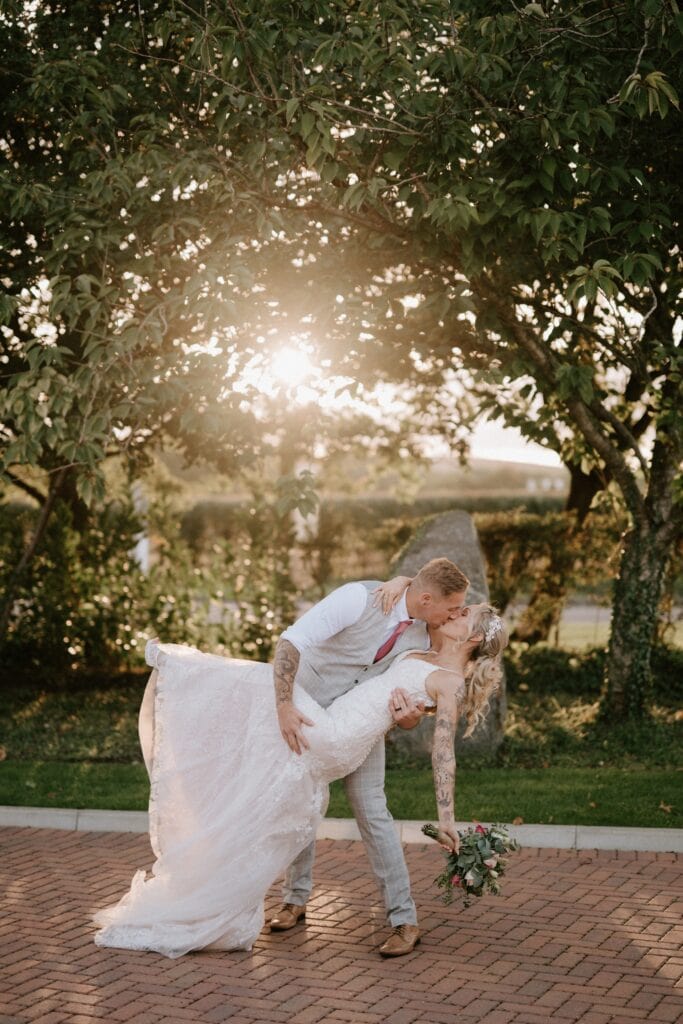 In the picturesque setting of Alfriston Gardens, a groom in a light gray suit dips his bride in a lace wedding dress for a kiss under a tree, with the sun shining through the branches. The bride holds a bouquet as they stand surrounded by a garden and stone path, capturing their perfect wedding moment. Image by Pearce Wedding Photography.