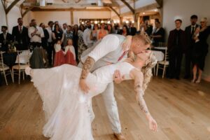 At Alfriston Gardens, a groom in a light grey suit passionately dips and kisses his bride, who is wearing a lace wedding dress, on the wooden dance floor. Guests in formal attire watch and smile, surrounding the couple in the warmly lit venue. Image by Pearce Wedding Photography.
