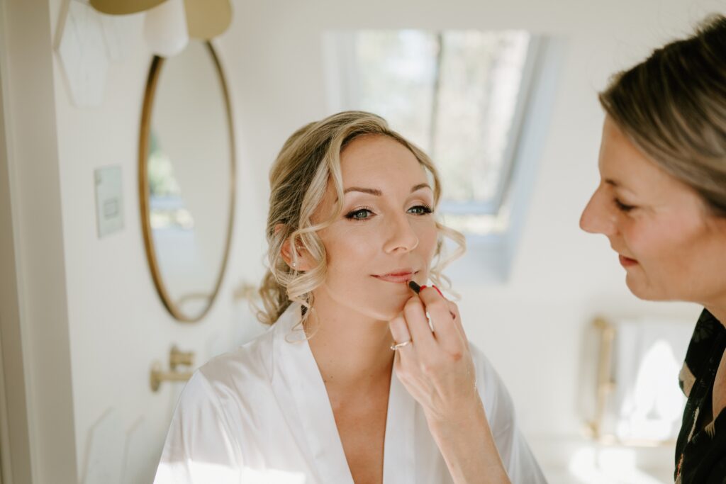 A woman getting ready for a wedding, wearing a white robe, sits in front of a round mirror while another person applies lipstick. The setting is bright and airy, reminiscent of Alfriston Gardens, with a window in the background. Image by Pearce Wedding Photography.