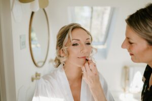 A woman getting ready for a wedding, wearing a white robe, sits in front of a round mirror while another person applies lipstick. The setting is bright and airy, reminiscent of Alfriston Gardens, with a window in the background. Image by Pearce Wedding Photography.