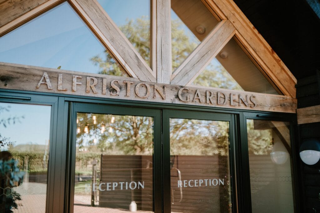 The entrance to the building features elegant wooden beams and glass doors, welcoming guests to "Alfriston Gardens." The word "Reception" is gracefully etched on the glass, perfect for a picturesque wedding venue. Tree and lantern reflections dance softly across the transparent surface. Image by Pearce Wedding Photography.