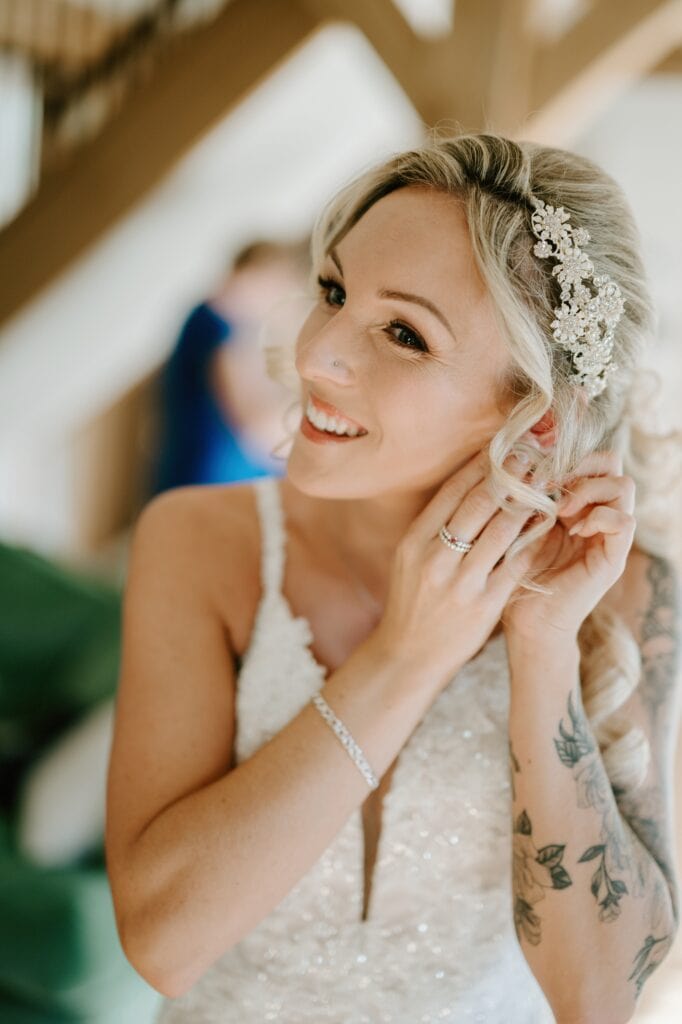 A smiling bride with blonde hair adjusts her earrings amidst the enchanting Alfriston Gardens. She wears a sparkly white dress and a decorative hairpiece. Visible tattoos adorn her arm, adding a unique charm, while the blurred background enhances this magical wedding moment. Image by Pearce Wedding Photography.