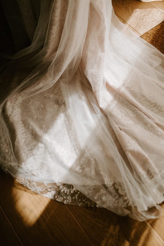 A close-up of a flowing, light-colored, textured fabric, possibly a bridal gown, draped elegantly over a wooden floor at Alfriston Gardens. The wedding fabric catches sunlight, highlighting its intricate patterns and delicate sheen. Image by Pearce Wedding Photography.