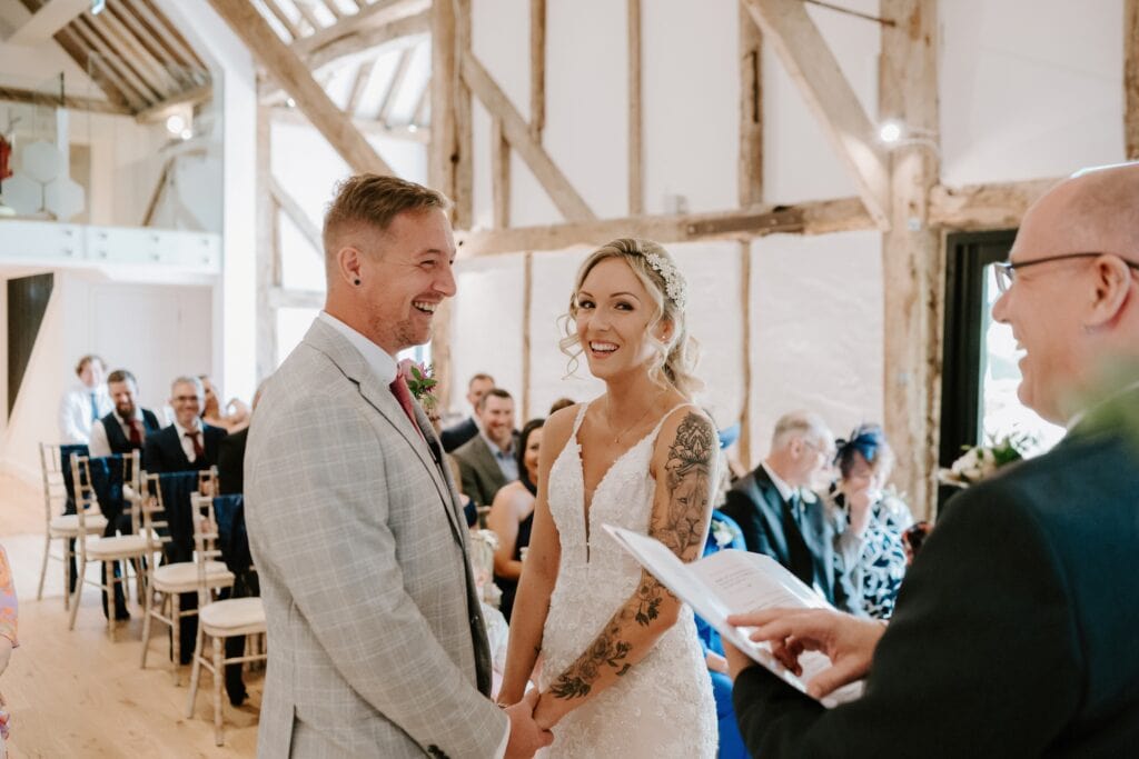 A couple stands smiling at each other during a wedding ceremony at the charming Alfriston Gardens, surrounded by rustic wooden beams. The groom wears a light gray suit, and the bride dons a white lace dress. Guests are seated in the background, and the officiant holds a booklet. Image by Pearce Wedding Photography.