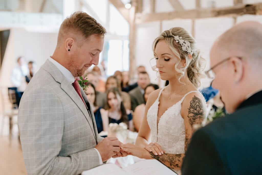 Amidst the enchanting setting of Alfriston Gardens, a groom in a light gray suit places a ring on the bride's finger during their wedding ceremony. The bride, adorned in a white lace dress and floral headpiece, stands opposite him, as an officiant presides and guests are seated in the background. Image by Pearce Wedding Photography.