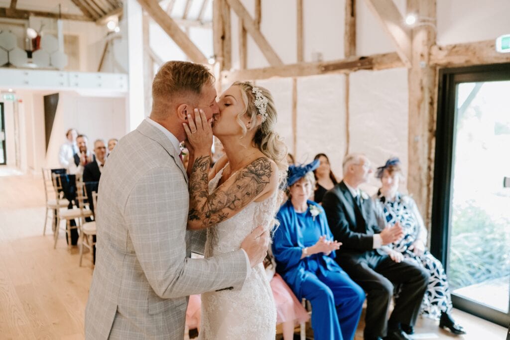 Amidst the enchanting charm of a rustic Alfriston Gardens venue, a bride and groom share a tender kiss during their wedding ceremony. The bride dazzles in a lace gown while the groom impresses in a light gray suit. Guests in formal attire watch and applaud the loving couple. Image by Pearce Wedding Photography.