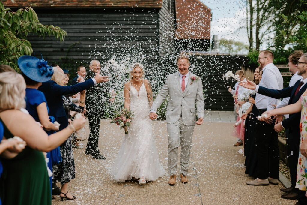 A bride and groom walk hand in hand down a path at Alfriston Gardens, surrounded by guests throwing white confetti. The bride wears a white gown and holds a bouquet, while the groom is in a light gray suit with a pink tie. Guests are dressed in colorful outfits, completing the wedding's vibrant atmosphere. Image by Pearce Wedding Photography.