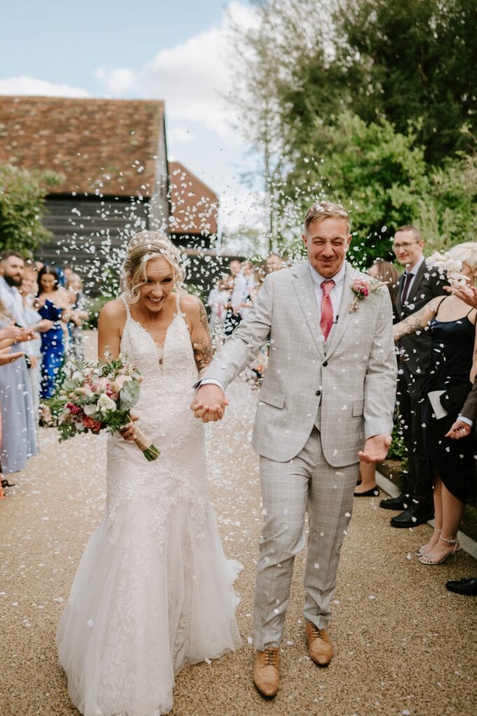 In the enchanting Alfriston Gardens, a bride and groom are walking down an outdoor aisle, holding hands and smiling. The bride stuns in a white dress with a bouquet, while the groom complements her in a light gray suit. Guests cheer and throw confetti, celebrating this magical wedding moment. Image by Pearce Wedding Photography.