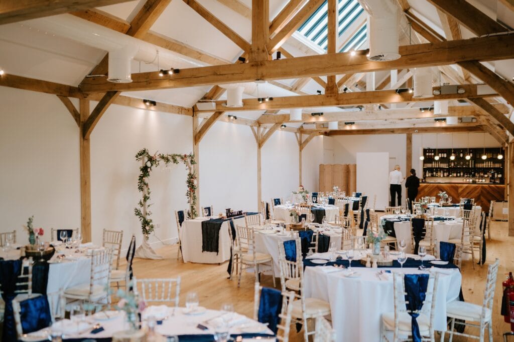 A spacious, rustic wedding venue in Alfriston Gardens, featuring exposed wooden beams and white walls. Round tables are elegantly set with white linens and navy accents. The room boasts a floral arch and a bar area at the far end, perfect for celebrating your special day. Image by Pearce Wedding Photography.