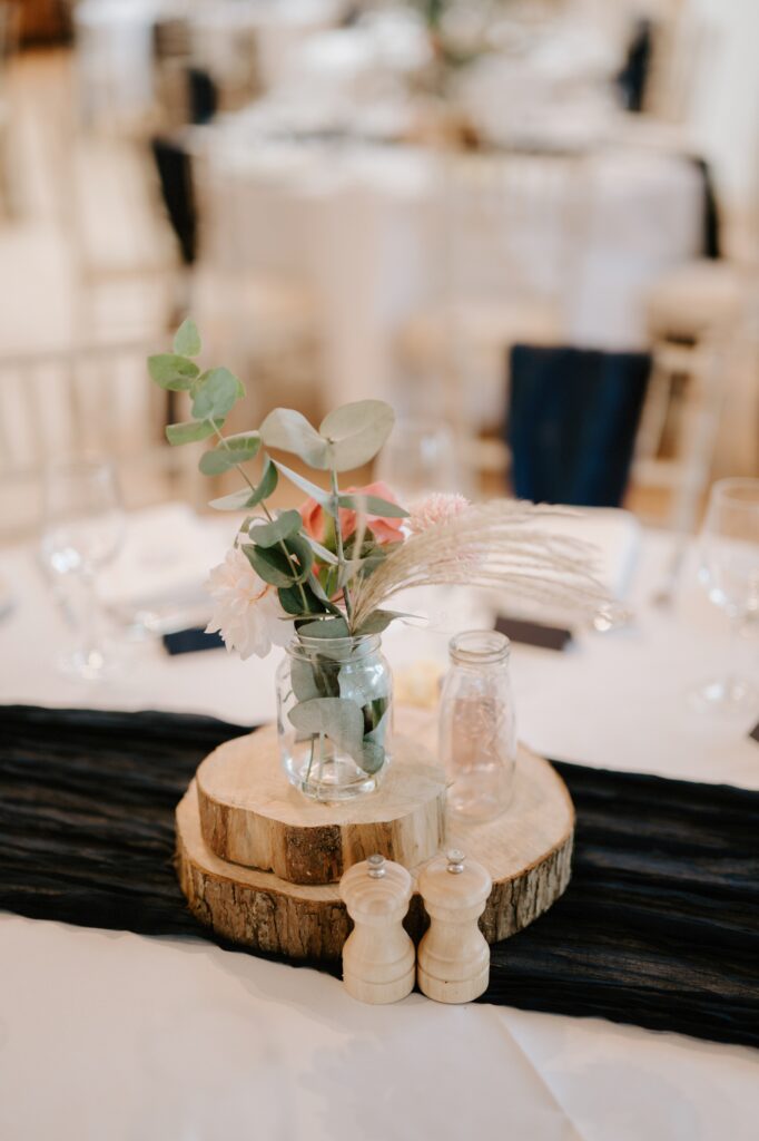A table centerpiece perfect for an Alfriston Gardens wedding features a small vase with eucalyptus leaves and pink flowers on wooden slices. Nearby, empty glass bottles and salt and pepper shakers rest elegantly on a dark runner. Image by Pearce Wedding Photography.