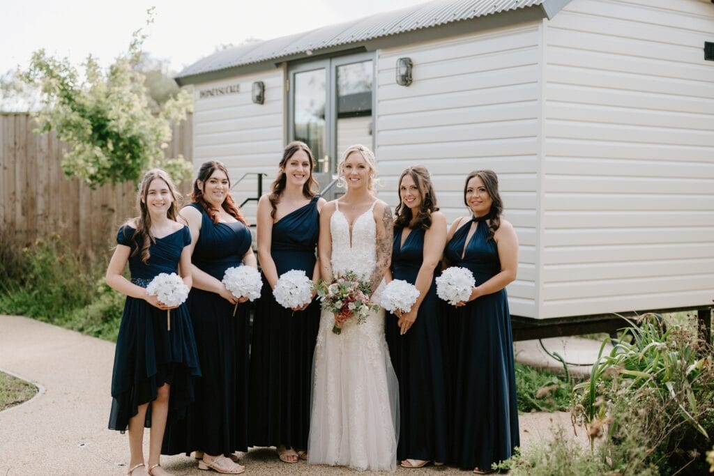 A bride in a white dress stands with five bridesmaids wearing dark blue dresses, each holding white bouquets. They pose gracefully on the garden path in front of a light-colored building at Alfriston Gardens, capturing a timeless wedding moment. Image by Pearce Wedding Photography.