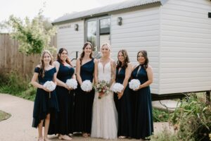 A bride in a white dress stands with five bridesmaids wearing dark blue dresses, each holding white bouquets. They pose gracefully on the garden path in front of a light-colored building at Alfriston Gardens, capturing a timeless wedding moment. Image by Pearce Wedding Photography.
