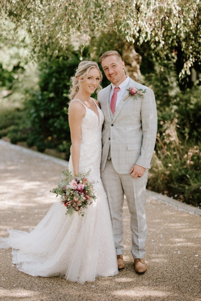 A couple poses outdoors on their wedding day at Alfriston Gardens. The bride wears a lace gown and holds a bouquet of pink and white flowers. The groom is in a light gray suit with a pink tie. They stand on a path surrounded by lush greenery and dappled sunlight. Image by Pearce Wedding Photography.