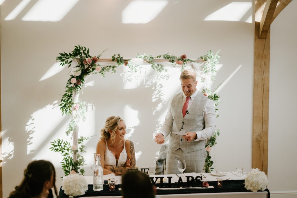 At their wedding in Alfriston Gardens, a groom stands with a microphone, smiling and holding notes. Beside him, the bride laughs. They sit at a decorated table adorned with greenery and floral arrangements, set before a backdrop featuring their names. Image by Pearce Wedding Photography.