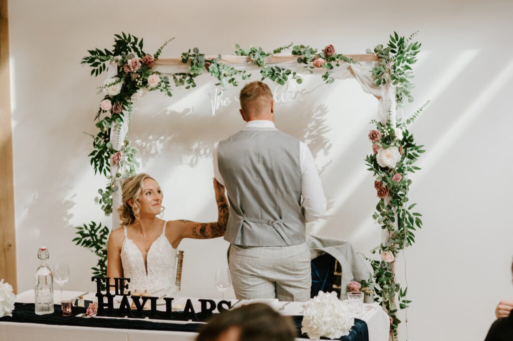 A bride in a white dress sits at a decorated table at Alfriston Gardens, her back to a groom in a gray suit who stands beside her. Under a floral arch with greenery and roses, a decorative sign on the table reads, "The Taylors," capturing the enchanting essence of their wedding day. Image by Pearce Wedding Photography.