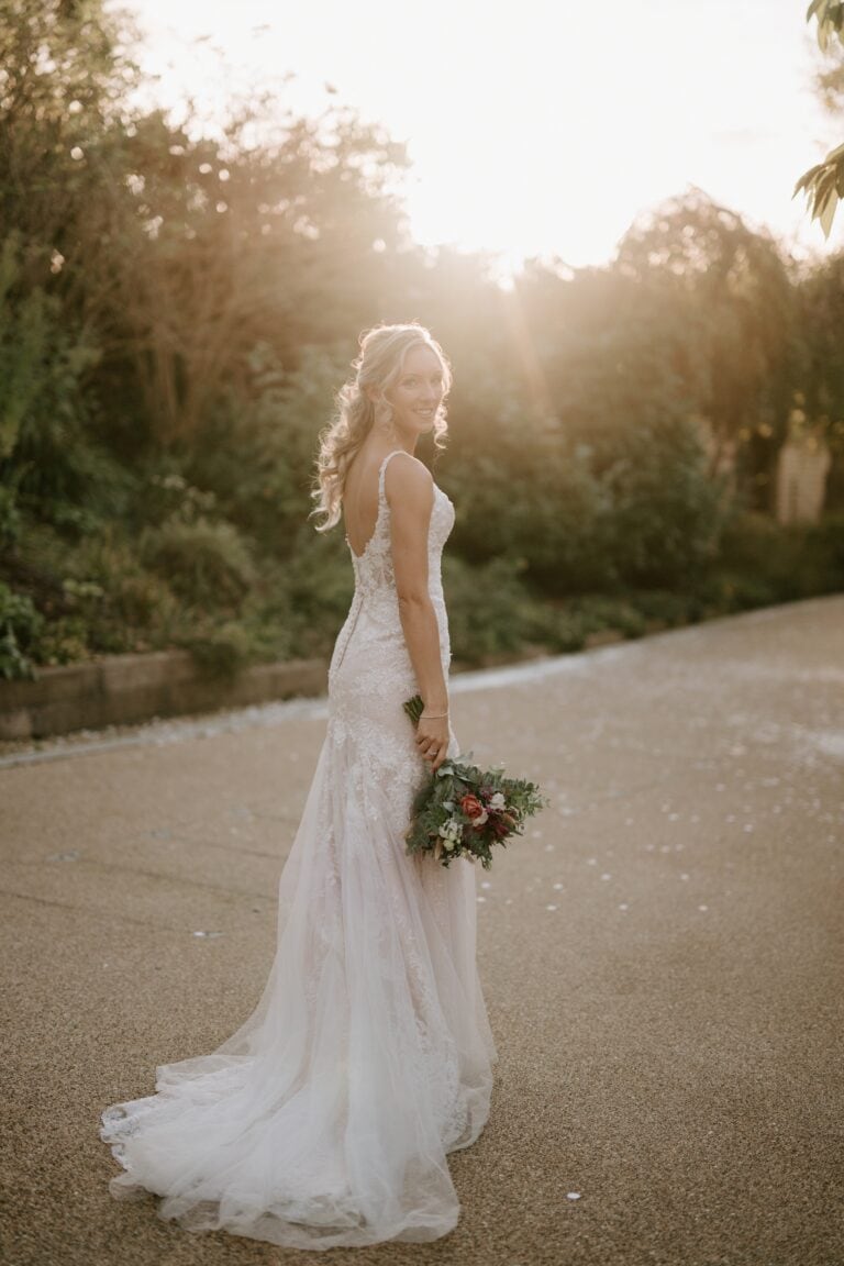A bride in a lace gown stands on the pathway of Alfriston Gardens, holding a bouquet of flowers. She looks over her shoulder with a smile, illuminated by the warm glow of the setting sun as lush greenery envelops the enchanting wedding scene. Image by Pearce Wedding Photography.