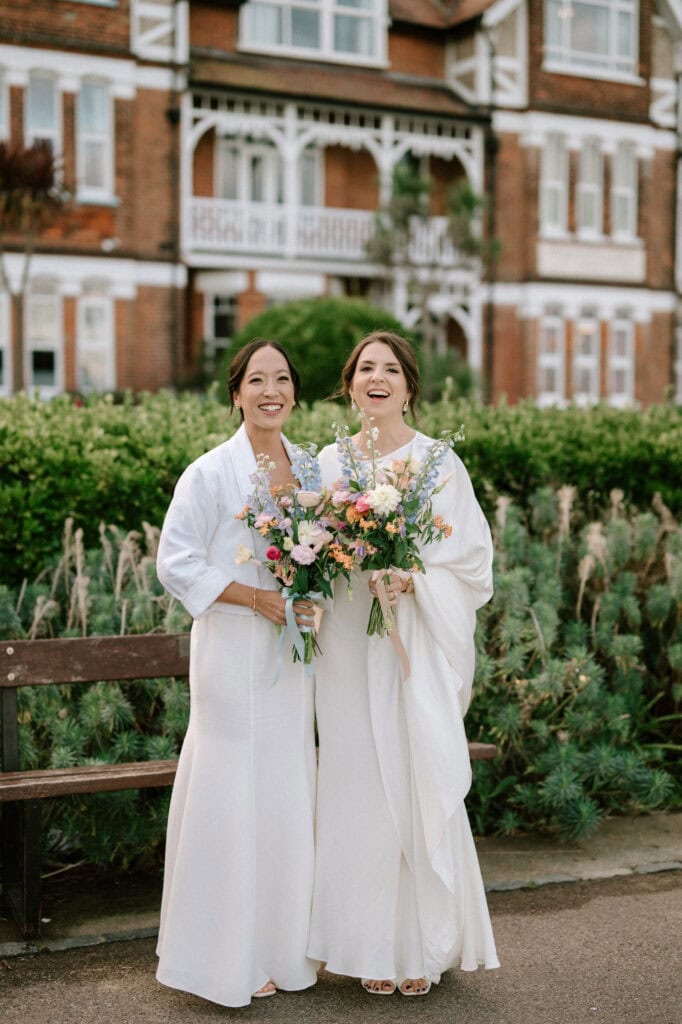 Two women in white dresses stand joyfully in front of a brick building with green hedges, holding colorful bouquets. Their smiles suggest the presence of a Kent wedding photographer capturing this special outdoor moment, possibly at a wedding or similar event. Image by Pearce Wedding Photography.