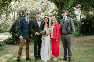 A stunning example of wedding photography, this group photo captures a bride in a white dress and bouquet, a groom in a green suit, and three others—two men in suits and a woman in a bright pink outfit—against the lush backdrop of trees and grass. Image by Pearce Wedding Photography.