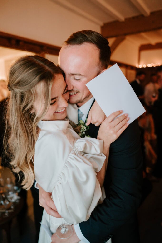 A joyful couple embraces warmly at a celebration, captured beautifully by a Kent wedding photographer. The woman smiles while holding a white card, and the man is dressed in a suit with a boutonniere on his lapel. Soft, ambient lighting and a blurred background create an intimate atmosphere. Image by Pearce Wedding Photography.