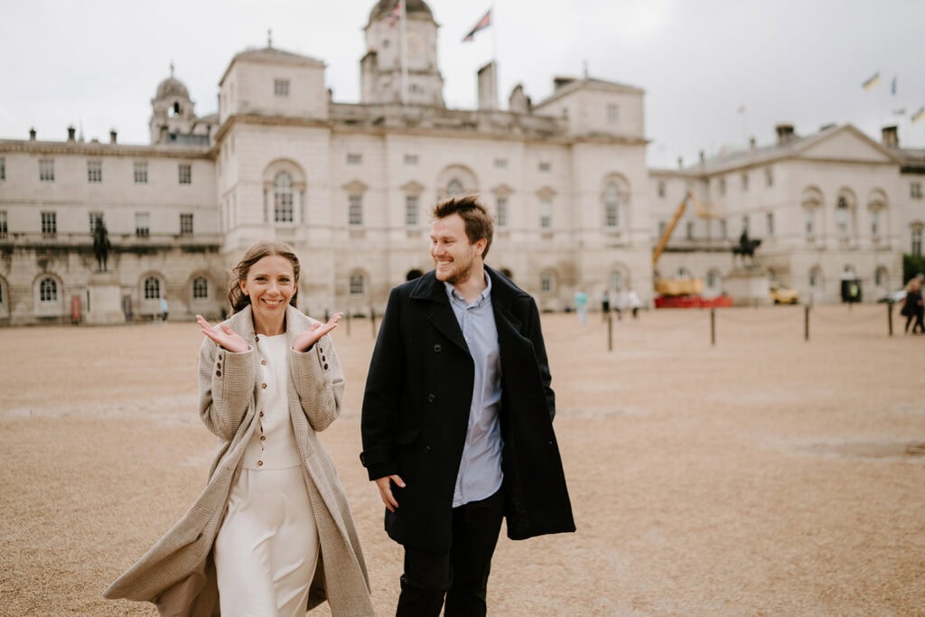 A smiling woman in a beige coat gestures happily with her hands, standing next to a man in a black coat. In this enchanting pre-wedding moment, they stand in an open area with a historical building behind them and a cloudy sky above. Image by Pearce Wedding Photography.