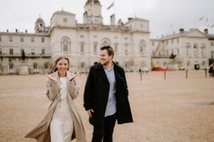 A smiling woman in a beige coat gestures happily with her hands, standing next to a man in a black coat. In this enchanting pre-wedding moment, they stand in an open area with a historical building behind them and a cloudy sky above. Image by Pearce Wedding Photography.