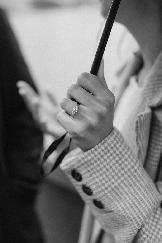 A black and white image captures a pre-wedding moment with a person holding an umbrella. The focus is on their hand, adorned with an engagement ring featuring a prominent oval-shaped stone. They wear a textured coat, its button detail clear, while the background remains blurred. Image by Pearce Wedding Photography.