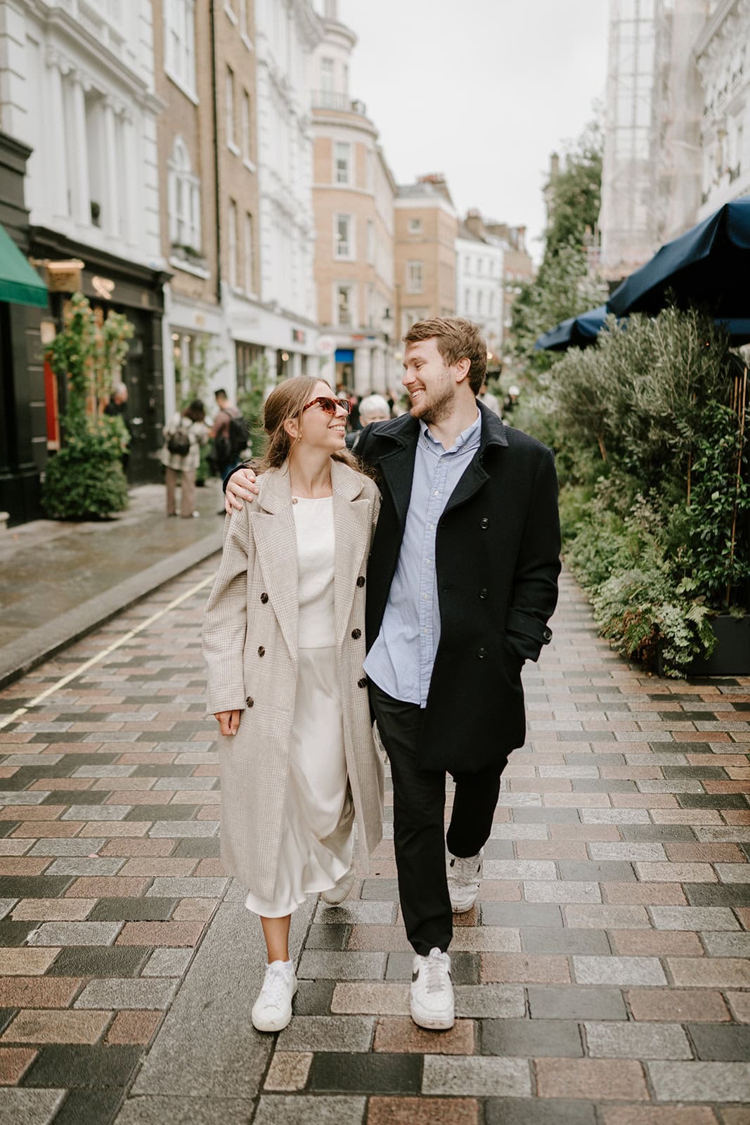 A smiling couple walks arm in arm down a cobblestone street, reminiscent of charming London pathways. The woman is wearing a long beige coat and sunglasses, while the man sports a dark coat and blue shirt. Surrounded by buildings and greenery on this cloudy day, it feels like the perfect pre-wedding stroll. Image by Pearce Wedding Photography.