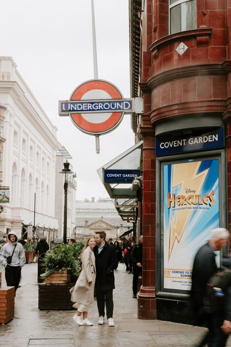 A couple, perhaps celebrating their pre-wedding bliss, stands on a street under a London Underground sign near Covent Garden. They embrace in front of a red-brick building featuring a Disney "Hercules" musical poster as pedestrians stroll by. The scene is lively and urban. Image by Pearce Wedding Photography.