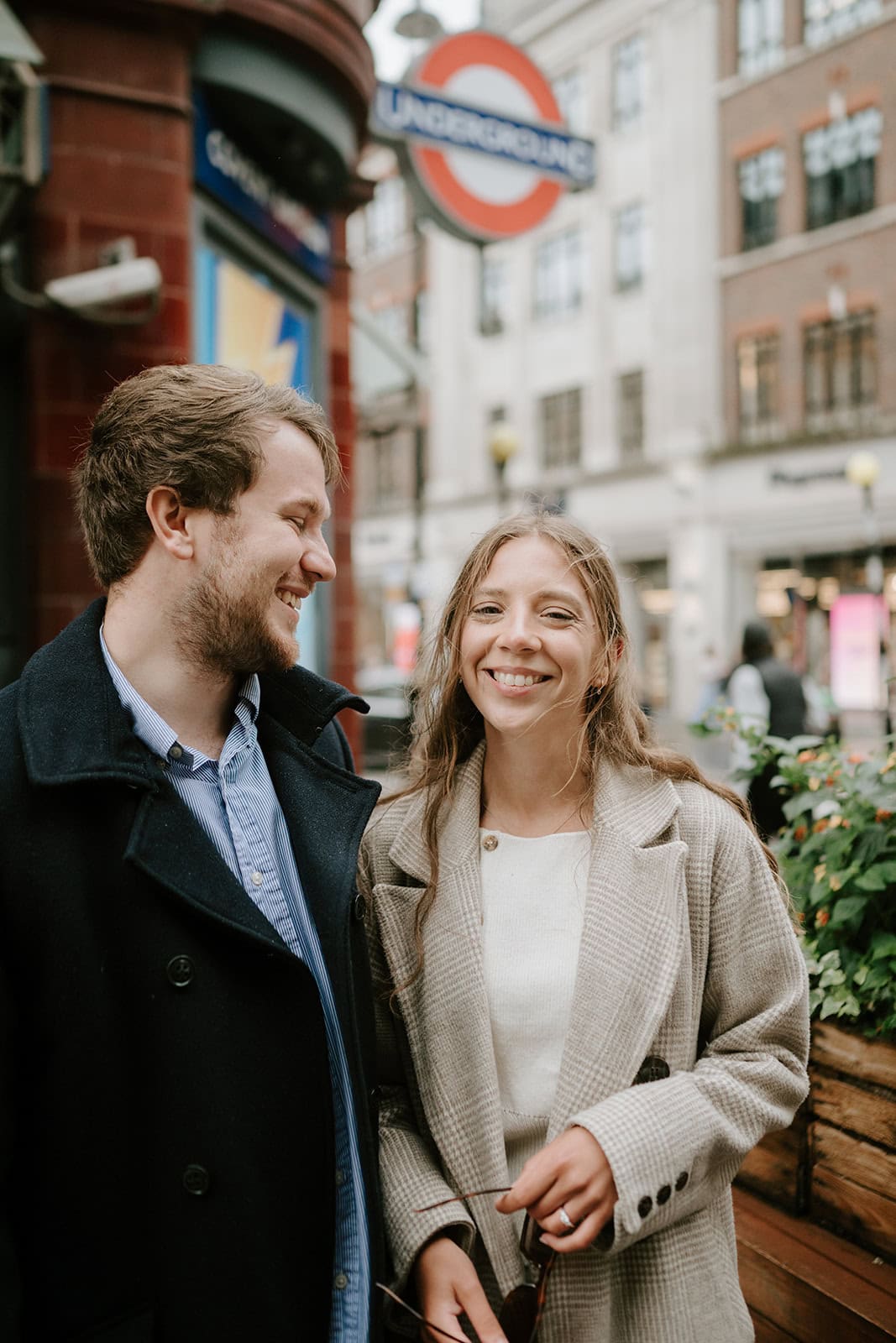 A man and a woman smiling at each other stand near a London Underground sign, reminiscent of engagement photography in Kent. The woman wears a light coat, and the man sports a dark one. They're outside next to potted plants, framed by city buildings in the backdrop. Image by Pearce Wedding Photography.