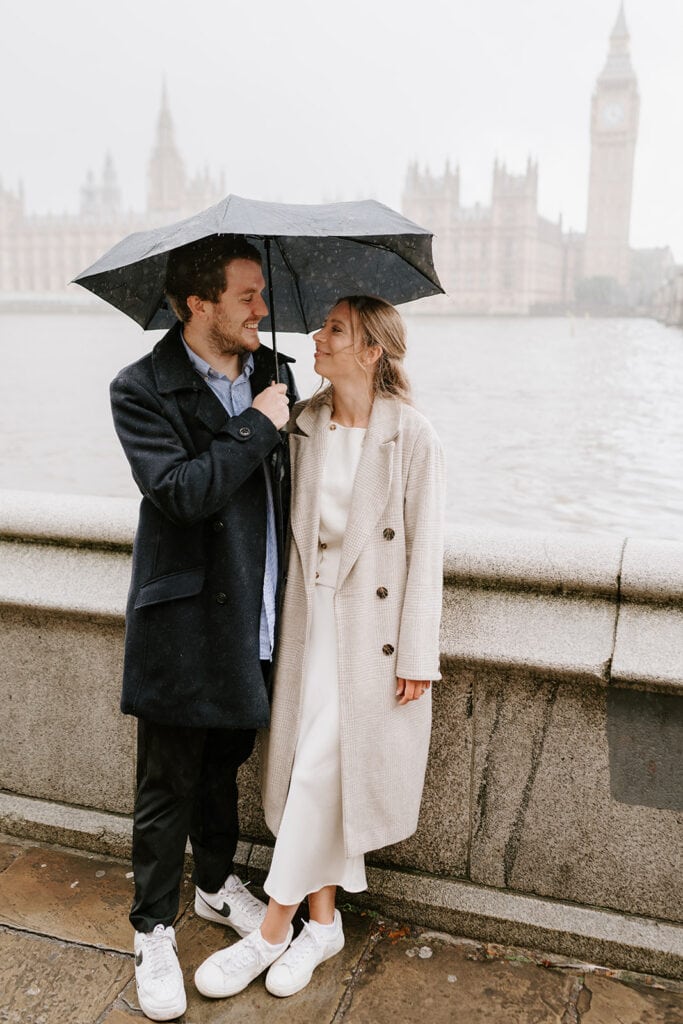 A couple stands under an umbrella in front of the Thames River with the Houses of Parliament and Big Ben in the background. The man wears a dark coat and the woman a light coat. Smiling, they gaze at each other on their rainy pre-wedding day. Image by Pearce Wedding Photography.