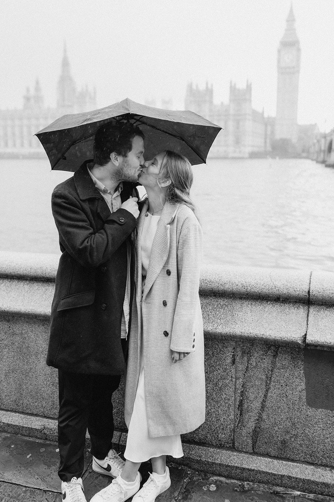 A cute couple enjoys a photoshoot in London, kissing under an umbrella on a rainy day. They stand by the river, with Big Ben and the Houses of Parliament as their backdrop, both wearing long coats and sneakers—a perfect romantic scene. Image by Pearce Wedding Photography.
