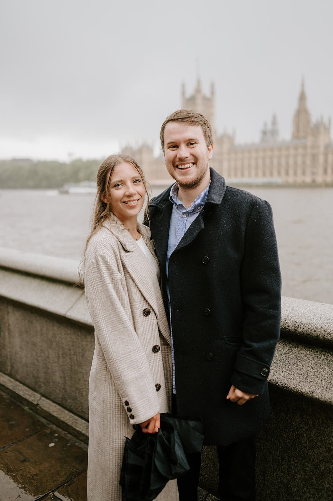 A cute couple stands smiling by the riverside on a cloudy day in London. They are both wearing coats, with the woman in a light coat and the man in a dark one. A historic building with towers looms charmingly across the water, enhancing their picturesque photoshoot setting. Image by Pearce Wedding Photography.