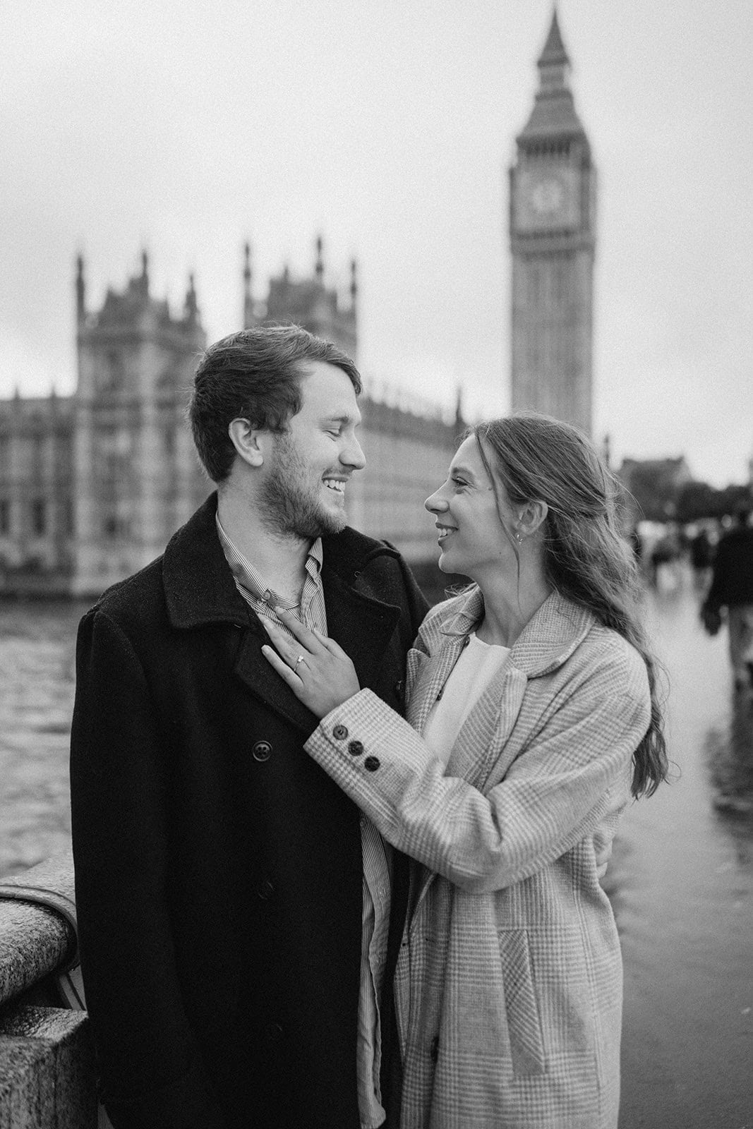 A cute couple photoshoot captures them smiling at each other in front of the Houses of Parliament and Big Ben in London. The scene is in black and white, with the iconic clock tower looming majestically on a cloudy day. Image by Pearce Wedding Photography.