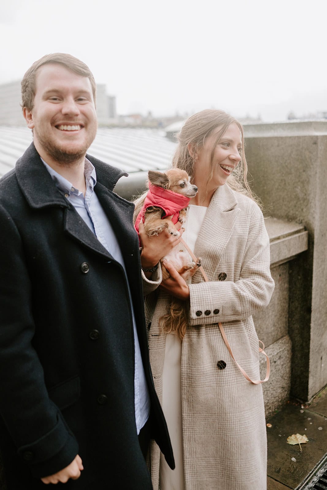A smiling couple poses for a cute photoshoot outside, holding a small dog in a pink outfit. The man sports a dark coat, the woman a light one. They're standing before a stone wall with London's iconic cityscape in the cloudy backdrop. Image by Pearce Wedding Photography.