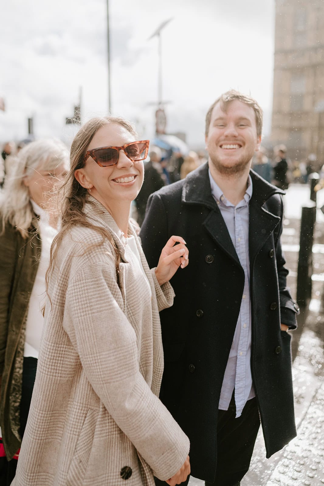 A woman in sunglasses and a man in a dark coat, perhaps celebrating a pre-wedding moment, are smiling and walking outside on a rainy Southwark day. The background reveals other people and blurred urban elements, while the bright light suggests a mix of rain and sunshine. Image by Pearce Wedding Photography.