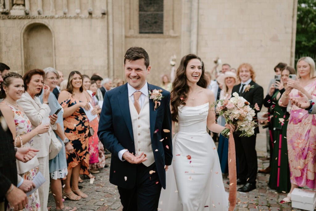 A bride and groom walk joyfully down a cobblestone path, surrounded by a cheering crowd. The bride holds a bouquet and wears a white dress, while the groom, captured beautifully by a Kent wedding photographer, wears a dark suit. Guests toss flower petals in celebration outside a historic building.