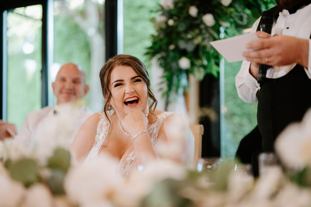 A smiling bride sits at a table, surrounded by floral arrangements, laughing as she rests her chin on her hand. Nearby, a person in formal attire speaks into a microphone. Lush greenery frames the joyful scene at The Old Kent Barn wedding. Image by Pearce Wedding Photography.