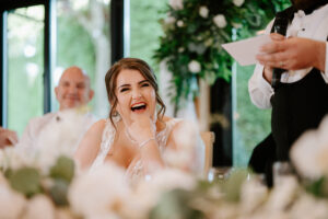 A smiling bride sits at a table, surrounded by floral arrangements, laughing as she rests her chin on her hand. Nearby, a person in formal attire speaks into a microphone. Lush greenery frames the joyful scene at The Old Kent Barn wedding. Image by Pearce Wedding Photography.
