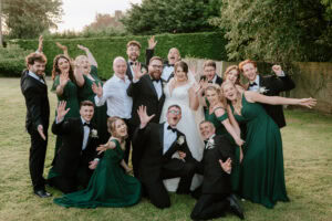 A joyful wedding party poses on the grassy lawn of The Old Kent Barn. The bride and groom stand at the center, surrounded by bridesmaids in green dresses and groomsmen in black suits. With arms raised and smiles wide, they celebrate this happy occasion. Image by Pearce Wedding Photography.