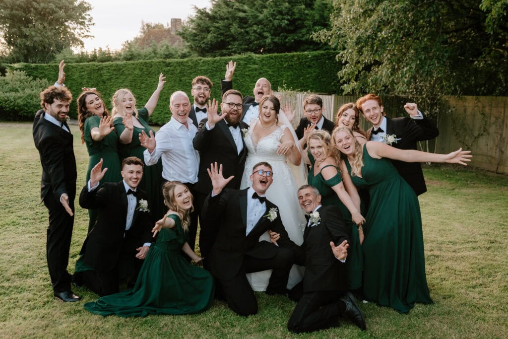 A joyful wedding party poses outdoors on a lawn at The Old Kent Barn, with the bride and groom at the center. The group, dressed in formal attire with green dresses and black suits, smiles and waves hands excitedly. Trees and a fence are in the background. Image by Pearce Wedding Photography.