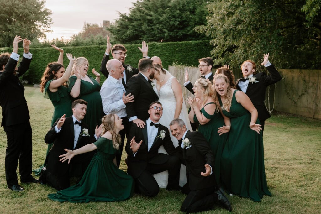 A joyful wedding party poses playfully outdoors at The Old Kent Barn, with the bride and groom kissing in the center. Bridesmaids in green dresses and groomsmen in black suits energetically gesture around them, all on a grassy lawn framed by towering trees. Image by Pearce Wedding Photography.