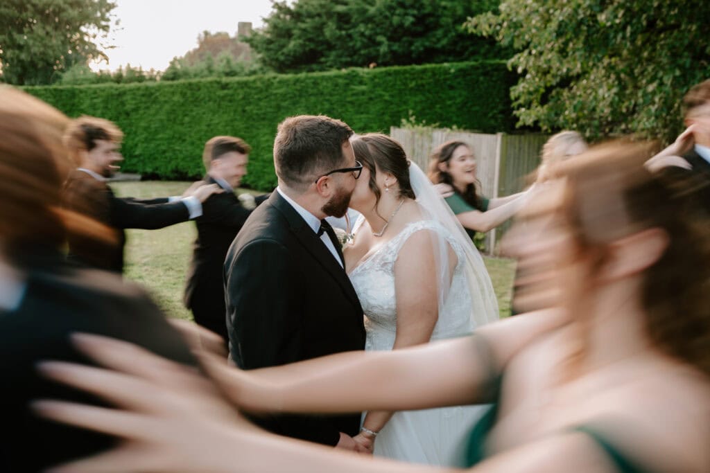 A bride and groom share a kiss outdoors at the Old Kent Barn, while a blurred group of people joyfully dance around them. The background is filled with greenery, and the sunlight softly highlights this enchanting wedding scene. Image by Pearce Wedding Photography.