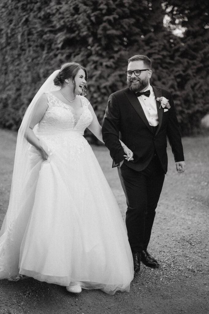 A couple in wedding attire joyfully walk hand in hand along a gravel path at The Old Kent Barn. The bride, in a flowing gown and veil, and the groom, in a tuxedo, smile at each other against a backdrop of tall, lush greenery. Image by Pearce Wedding Photography.