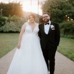 A bride in a white gown and a groom in a black tuxedo stand on a gravel path outside the Old Kent Barn. They are smiling with soft sunlight in the background, surrounded by lush greenery, capturing the perfect wedding moment. Image by Pearce Wedding Photography.