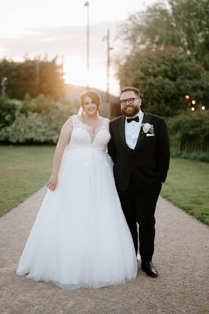 A bride in a white gown and a groom in a black tuxedo stand on a gravel path outside the Old Kent Barn. They are smiling with soft sunlight in the background, surrounded by lush greenery, capturing the perfect wedding moment. Image by Pearce Wedding Photography.