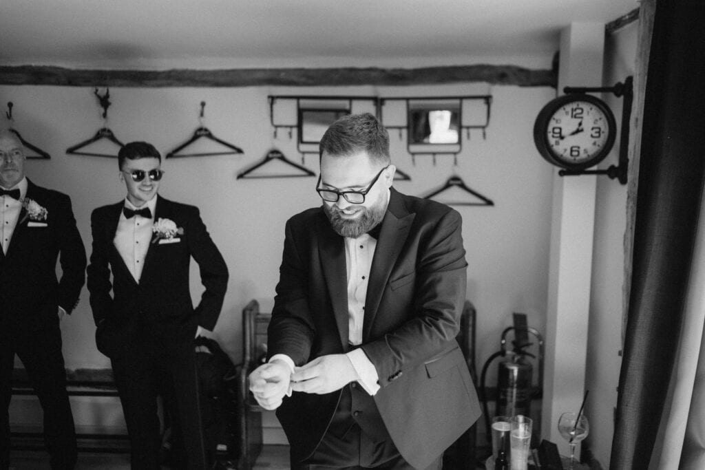A man in a suit adjusts his cufflinks in a room at The Old Kent Barn, preparing for the wedding. Two other men, also in suits, stand in the background; one wears sunglasses. Empty hangers line the wall, and an antique clock is mounted nearby. The atmosphere is formal and relaxed. Image by Pearce Wedding Photography.