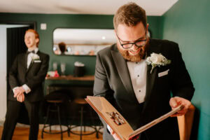 A groom in a black tuxedo and glasses joyfully reads a scrapbook at The Old Kent Barn. He stands in a room with a green wall and mirror, capturing the essence of the wedding day. Another man in a tuxedo laughs heartily near the bar area, adding to the joyous atmosphere. Image by Pearce Wedding Photography.